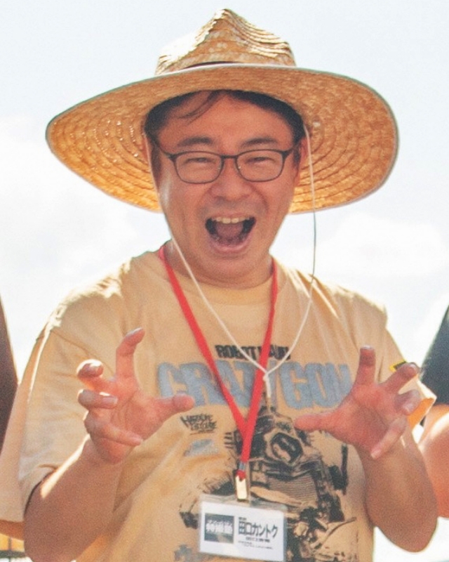 大きな麦わら帽子をかぶり胸元に名札を下げ両手の指を立てて怪獣のようなポーズを取る田口清隆塾長の写真