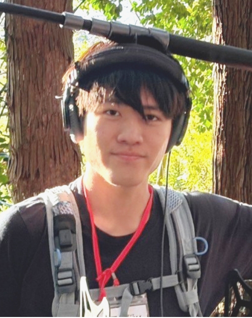 録音用ヘッドホンと機材ハーネスを装着し頭上のブームマイク持って立っている三門優介録音担当の写真