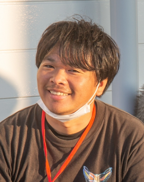 黒いTシャツに赤いストラップを下げ前方に体を向けて静かに座る渡邉聡メイキング担当の写真