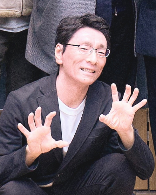 黒いジャケット姿のしゃがんだ姿勢で両手の指を大きく開き怪獣のようなポーズを取る島崎淳常任講師の写真