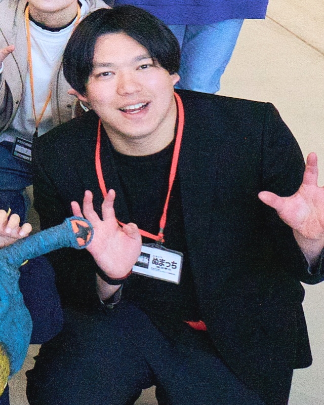 黒い服と名札を身につけ両手を広げ怪獣のようなポーズを取り小さな模型と並ぶ細沼孝之常任講師の写真