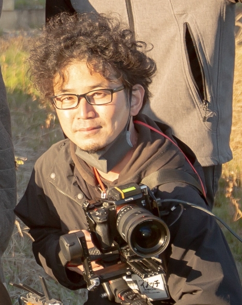 黒い上着を着て肩掛け装備の大型カメラを両手で構え撮影準備の姿勢を取る井野口功一撮影担当の写真