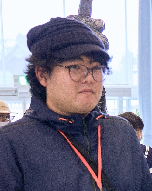 紺色の上着と赤いストラップを身につけ肩掛けバッグを下げて静かに立つ、名久井晨特撮美術補助の写真