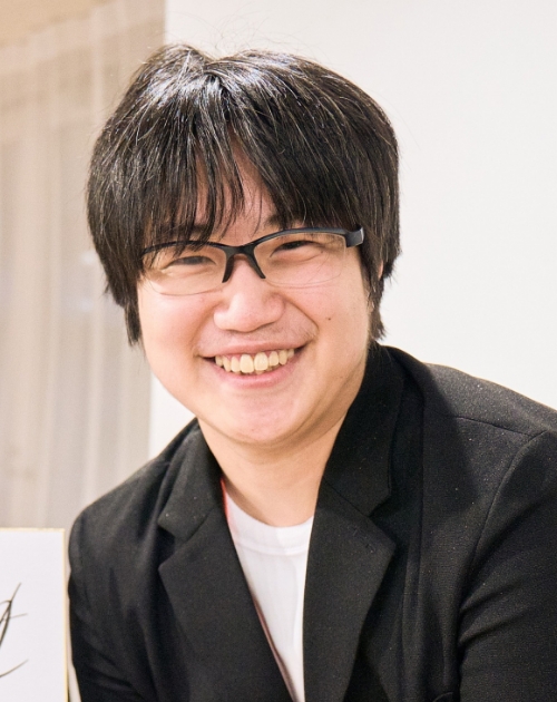 黒いジャケットに白いインナーを合わせ上体をわずかに傾けて前方を向き落ち着いた姿勢で座る鈴木優太特撮美術補助の写真