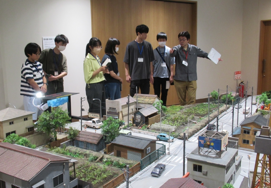 塾生たちが7人が並んで立ち精巧に作られた街並みの大型ジオラマを指差しながら構造や配置を熱心に確認して観察する様子の写真