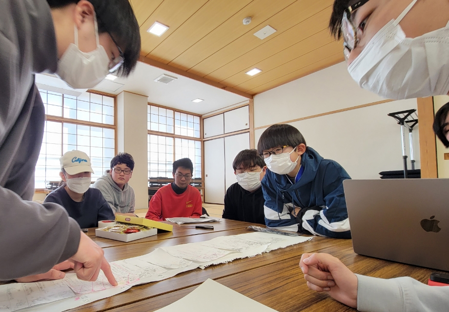 机上に広げた多数の資料やスケッチを囲み塾生が指示内容を見つめ意見交換しながら作業内容を確認している様子の写真