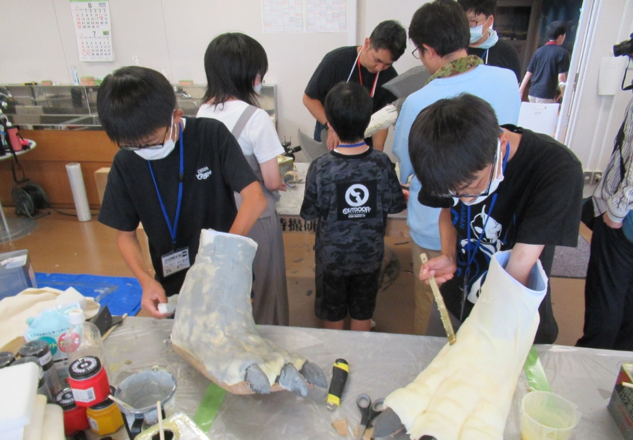 作業台に並ぶ巨大なモンスターの手型パーツに塾生の男性が塗装や成形作業を施し道具を使いながら丁寧に制作を進めている様子の写真