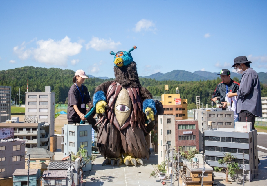 巨大な一つ目怪獣の着ぐるみが精巧なミニチュア都市の中央に立ち周囲でスタッフが撮影準備を進める様子の写真