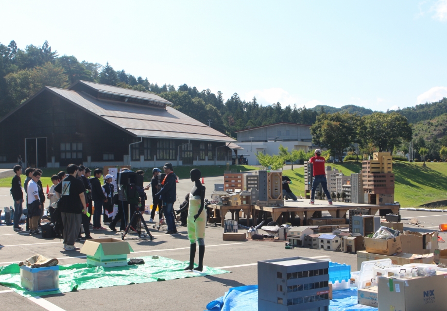 屋外の広場でスタッフと塾生がミニチュア建物を組み立て撮影用セットを調整しながら配置を確認している様子の写真