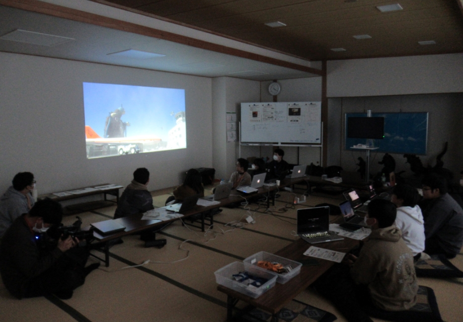暗い室内で塾生たちが床に座ってノートパソコンを広げ大型スクリーンに投影された特撮映像を見ながら編集内容や演出ポイントを真剣に確認している様子の写真