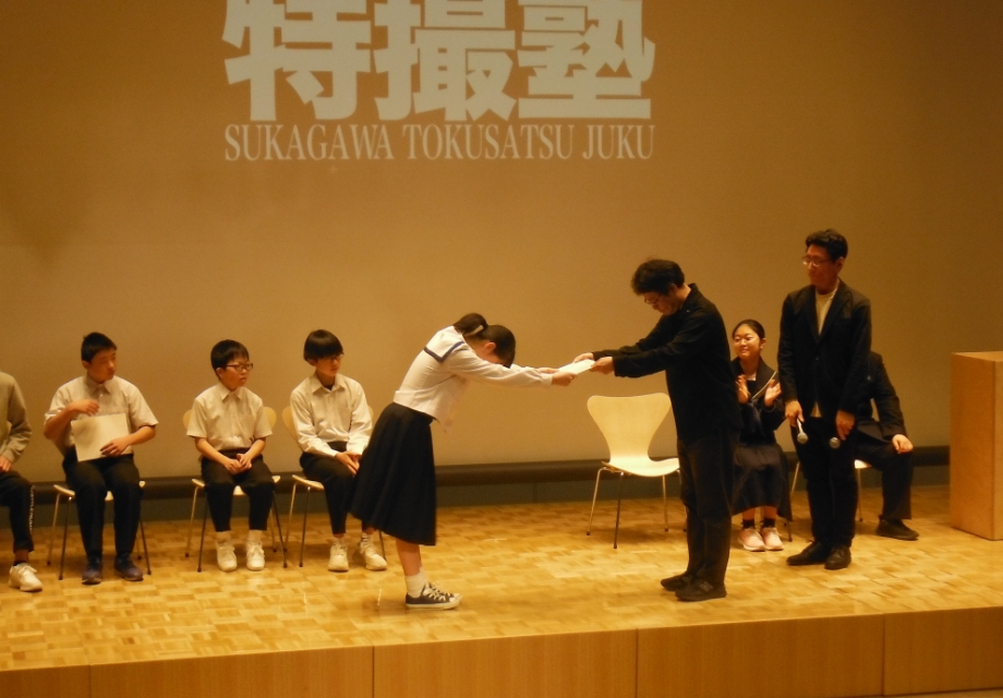 ステージ上で女子生徒が授与者に深く礼をしつつ証書を受け取り周囲の参加者が静かに見守る様子の写真