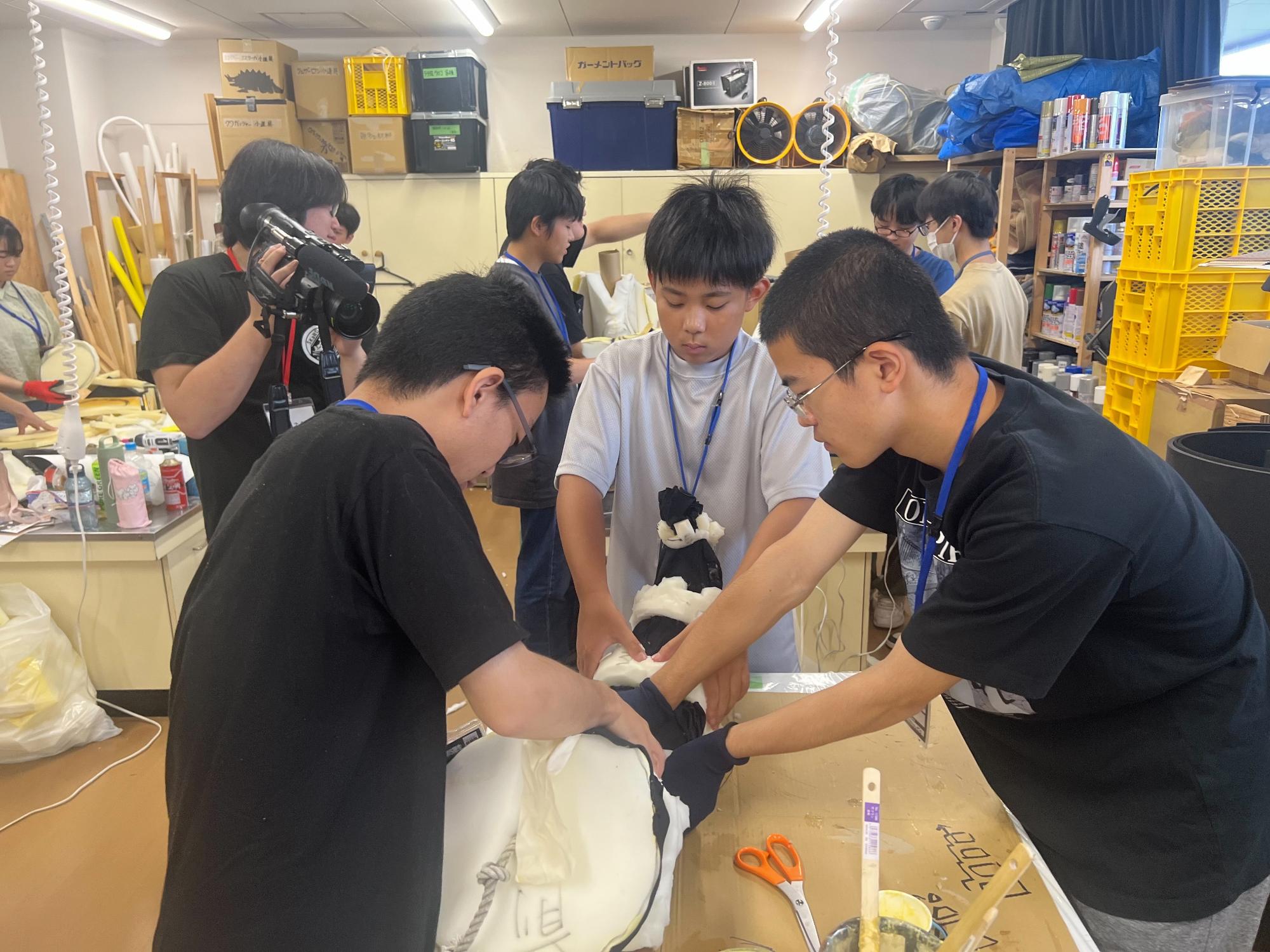 すかがわ特撮塾の活動で塾生たちが宇宙人の着ぐるみを制作している様子。写真ではしっぽの造形作業で数人が協力して接着作業をしている。