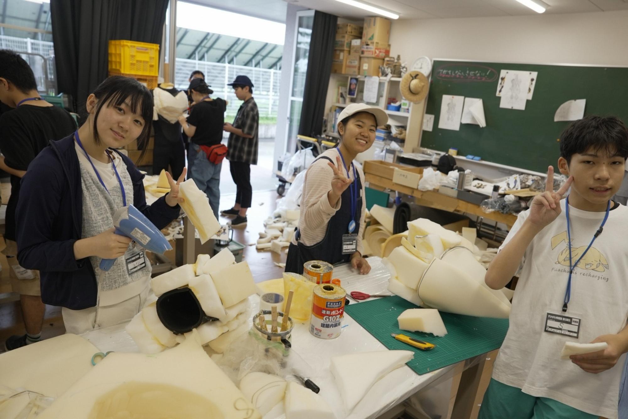 すかがわ特撮塾の活動で塾生たちが宇宙人の着ぐるみを制作している様子。手のパーツの造形作業の合間にカメラに向けてポーズをとっている。