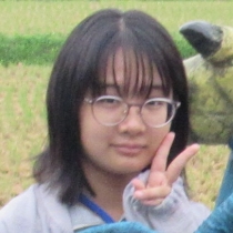 肩丈の髪の女性が眼鏡越しに視線を向けつつ片手でピースサインをしている様子の写真