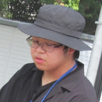 黒い帽子と濃色の上着を身につけた男性が斜め向きに立ち胸元にストラップを下げている様子の写真