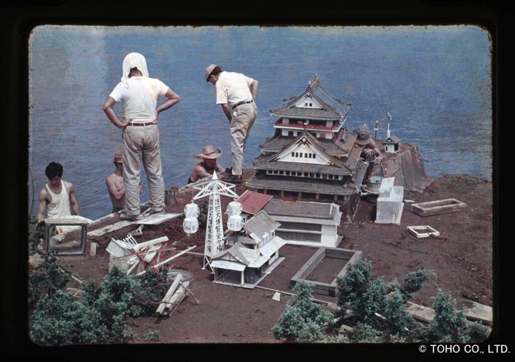 水辺に設置された精巧な日本城や城下町の巨大ミニチュアセットの周囲で、白い服を着たスタッフたちが配置の確認や細部の調整を行い、特撮映画の撮影準備を慎重に進めている記録を修正した写真