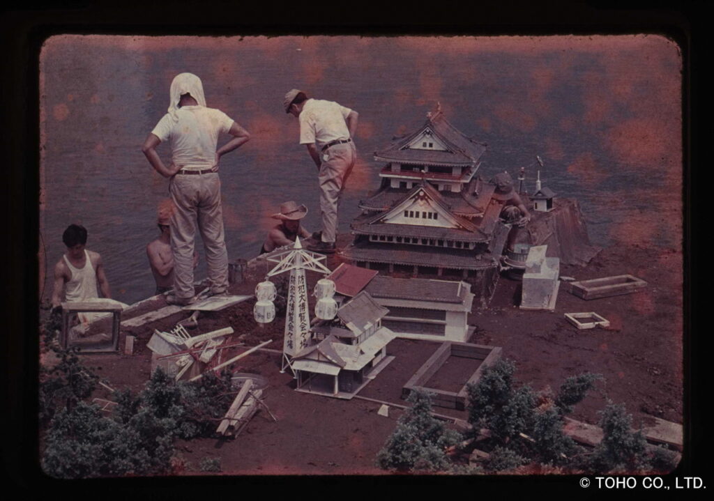 水辺に設置された精巧な日本城や城下町の巨大ミニチュアセットの周囲で、白い服を着たスタッフたちが配置の確認や細部の調整を行い、特撮映画の撮影準備を慎重に進めている記録が変色していることが分かる写真