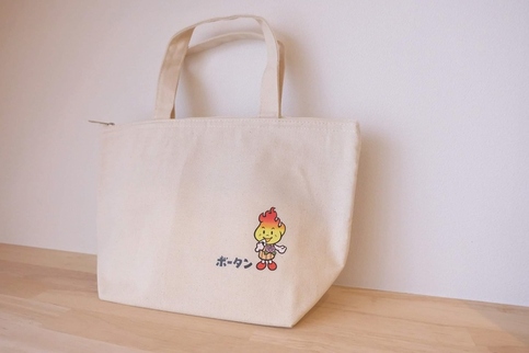 表面右下に、炎をモチーフにしたキャラクターボータンのイラストが描かれた、生成り色のランチバッグの写真