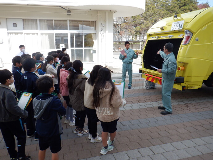 明るい屋外の校舎前で、作業服を着た職員が黄色いごみ収集車の後方にある投入口を指差しながら、集まった小学生たちにごみの収集の仕組みや分別の重要性について分かりやすく解説している様子を捉えた写真