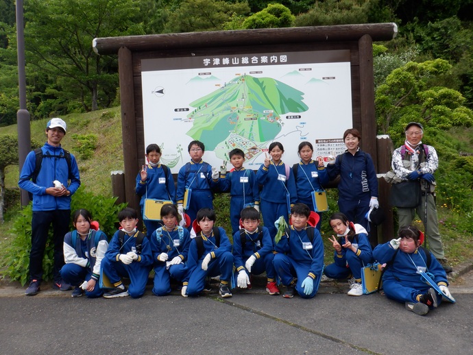 宇津峰山総合案内図という大きな看板の前に集まり、お揃いの青いジャージを着てVサインをする小学生たちと、それを引率する大人たちが緑豊かな自然を背景に並んでポーズを決めている集合写真
