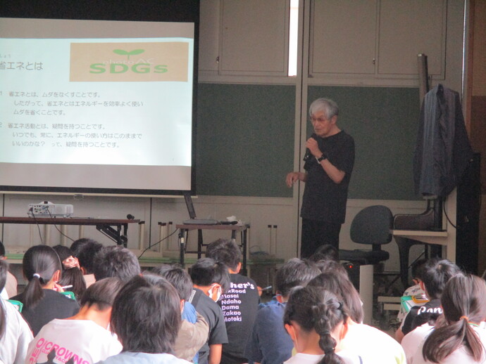 前方に映し出された「省エネとは」や「SDGs」という文字を含むスライドを背に、マイクを持って解説する男性と、その話を真剣に聞き入る大勢の子供たちの後ろ姿を捉えたESD環境教育の様子の写真