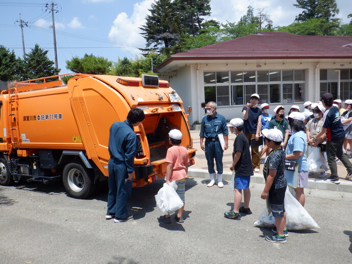ゴミ収集車を前に、ゴミ袋を手にした子供たちが並び、作業服を着た職員の方からゴミの収集や分別についての説明を熱心に聞いているESD環境教育の様子の写真