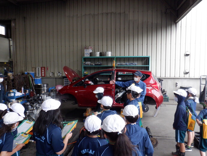 ボンネットやドアが開いた赤い乗用車の前に立ち身振り手振りで説明する作業服の男性を、白い帽子姿の小学生たちがメモを取り熱心に耳を傾けている様子の写真