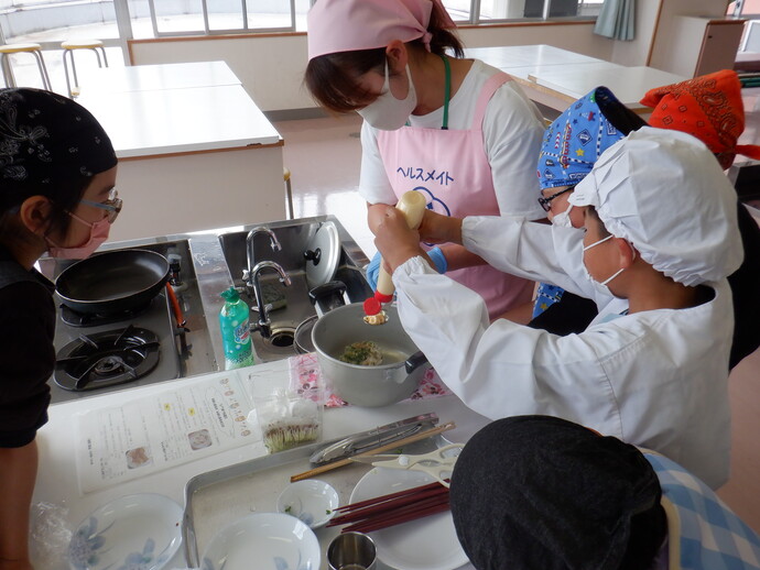 ピンクのエプロンをつけた指導員に見守られながら、バンダナや割烹着を身にまとった子供たちが協力して鍋の中にマヨネーズを絞り入れ、真剣な表情で料理を仕上げている様子の写真