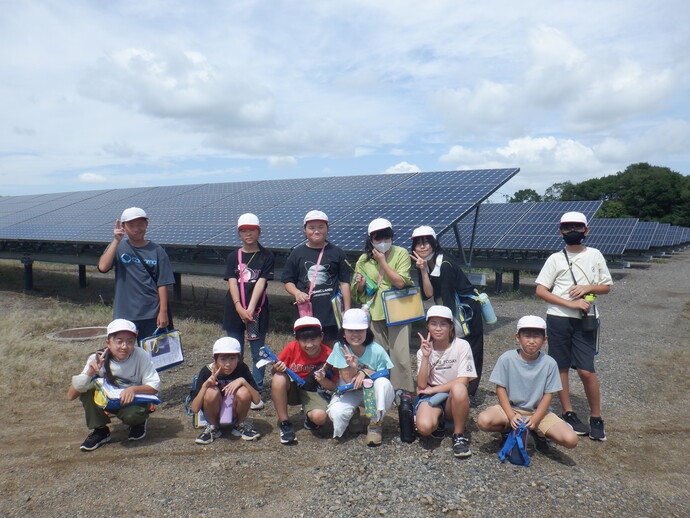 屋外の太陽光発電施設で、複数の人物が白い帽子をかぶり、ソーラーパネルの前に並んで立ったりしゃがんだりしており、手には水筒やバッグを持っている様子が見える写真