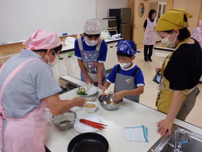 調理室のテーブルで、エプロンと三角巾を着けた複数の人物が料理の準備をしており、中央では子どもがボウルに卵を割り入れ、周囲には野菜や調味料、赤い箸、皿、布巾などの調理道具が並んでいる様子を写した写真