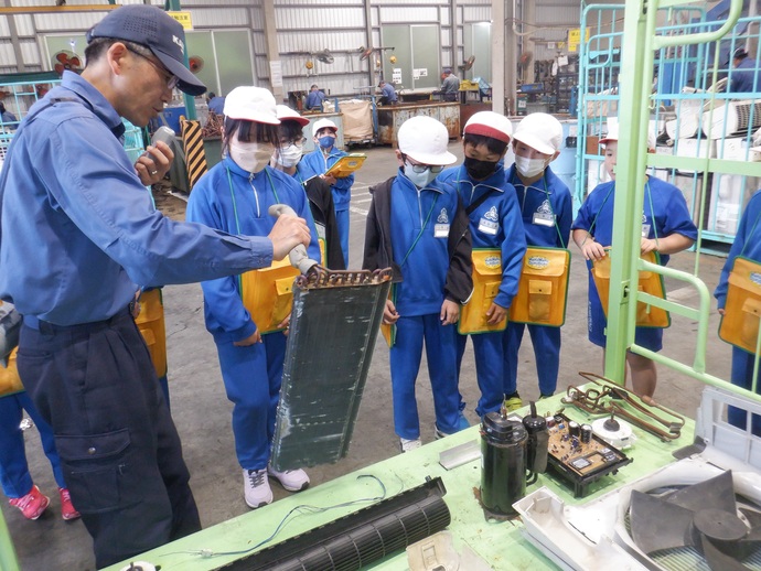 青い作業服を着た男性が、工場見学に訪れた小学生たちに向けてエアコンの内部パーツである熱交換器を手に持ちながら、リサイクルや製品の構造についてマイクを使って熱心に説明している様子を至近距離で捉えた写真