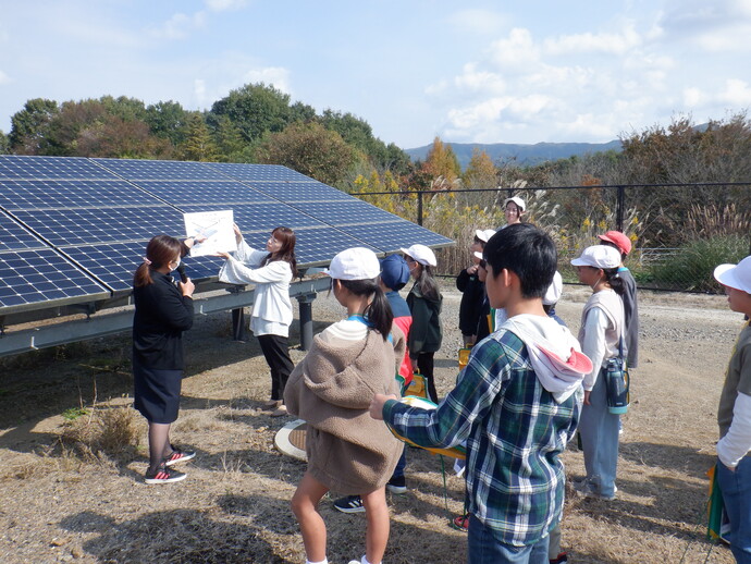 晴天の下でソーラーパネルの前に集まった小学生たちが、パネルの仕組みについて解説する女性の説明を熱心に聞きながら、再生可能エネルギーや環境問題について学外学習を行っている様子を捉えた写真