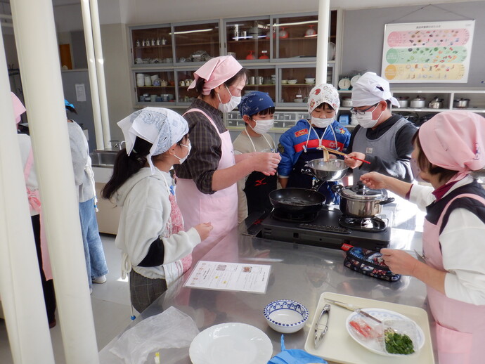 清潔感のある調理実習室で、エプロンと三角巾を着用した講師や小学生たちがコンロの前に集まり、鍋の中の状態を確認しながら調理の工程や食材の変化についての実践的な学びを深めている様子を捉えた写真