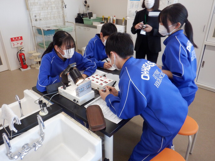理科室のような実験室で、お揃いの青いジャージを着た中学生たちが、机の上に置かれた小型の住宅模型やライト、測定機器を使いながら、太陽光発電や断熱効果に関する実験に真剣な表情で取り組んでいる様子を捉えた写真