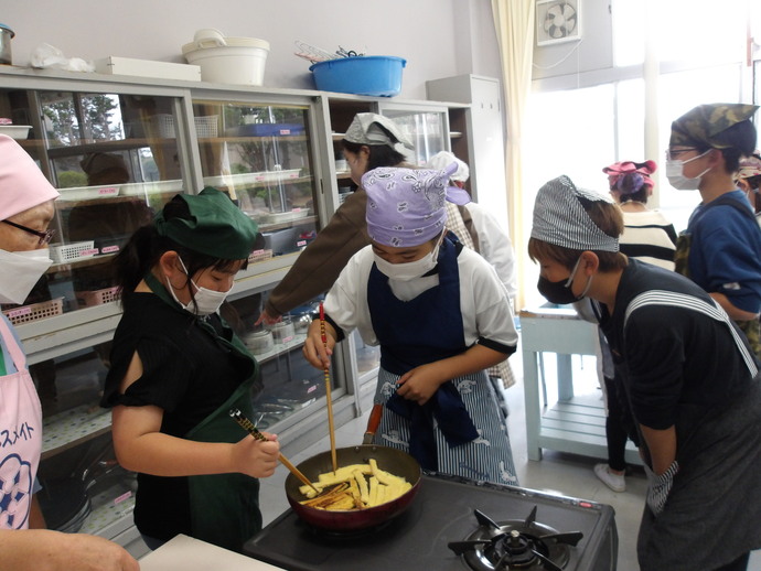 調理室でエプロンと三角巾を着けた人々が、ガスコンロの上で赤い鍋を使い、パンのみみフレンチトーストを菜箸で調理している様子の写真