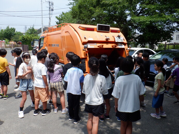 学校の校庭らしき場所で多数の子どもがオレンジ色のごみ収集車の後部を囲み、仕組みを見学している様子の写真