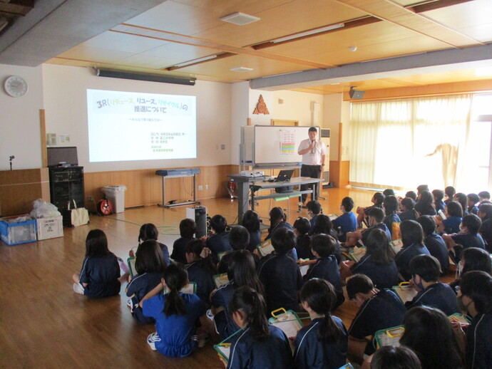 前方のスクリーンに映し出された「3R(リデュース、リユース、リサイクル)の推進について」というスライドを、床に座った大勢の子供たちが真剣に眺めながら講師の説明を聞いている、ESD環境教育の様子の写真