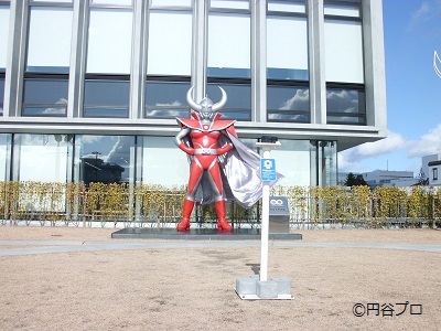 近代的なビルの前に立つ赤いスーツに銀色のマントを羽織り、大きな角を携え両手を腰に当てて堂々と佇むウルトラの父の等身大立像と、その前に設置されたカメラスタンドを写した写真