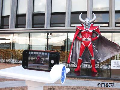 屋外に設置されたウルトラの父の立像の手前の撮影用スタンドスマートフォンが置かれ、ウルトラの父の立像が画面に映っている写真