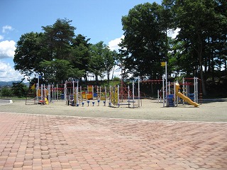 緑豊かな木々の手前に広がる砂地エリアに設置された、黄色い滑り台やジャングルジム、網状のアスレチックなどが組み合わさったカラフルな複合遊具の写真