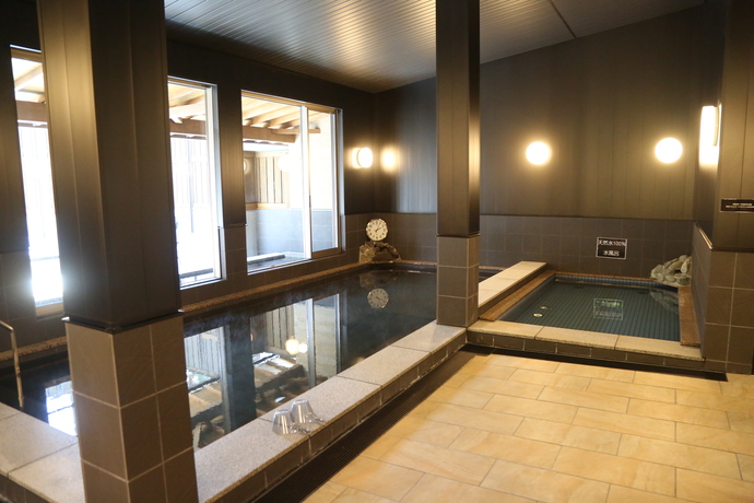 シックで落ち着いた黒い壁面に温かみのある丸い照明が灯り、石造りの広々とした大浴槽と小さめの浴槽が並び、大きな窓からは明るい光が差し込み清潔感あふれる浴場内を捉えた写真