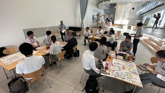 天井の高い開放的な会場で複数のグループに分かれて着席し、マイクを持って話す男性の方を向いて話を聞いている参加者たちの様子を、手前のテーブルに置かれた手書きの模造紙越しに俯瞰で撮影した第11回会議の様子写真
