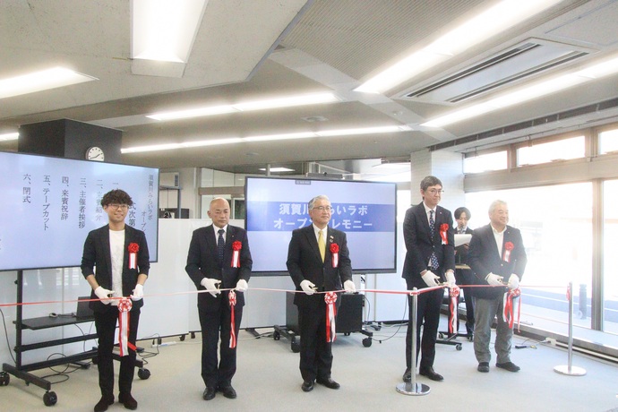 須賀川みらいラボのオープンセレモニー会場にて、スーツを着て胸に赤い花をつけた5人の男性関係者が横一列に並び、白手袋をしてハサミを持ち、記念のテープカットを行っている様子を撮影した写真