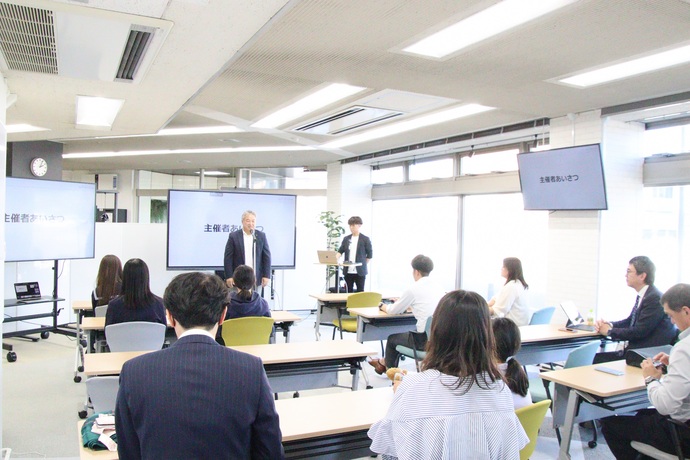 「主催者あいさつ」と表示されたモニターがある明るい会場の前方で、男性が参加者に向けてスピーチを行っており、長机に座った人々が背中を向けて静かにその話を聞いているワガママアワードの様子の写真