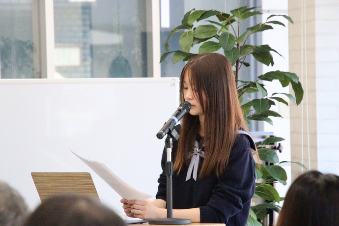 観葉植物が置かれた明るい室内で、紺色の制服を着たロングヘアの女性がスタンドマイクの前に立ち、手元の原稿に視線を落としながら真剣な表情で発表を行っている様子の写真