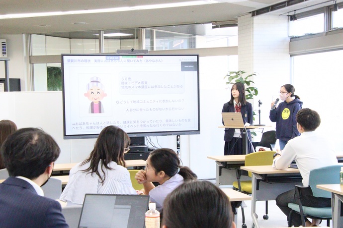 「須賀川市の現状」と書かれたスライドを表示したモニターの横で、制服とパーカー姿の二人の女子学生がマイクを使って発表し、手前の参加者が聞いている様子の写真
