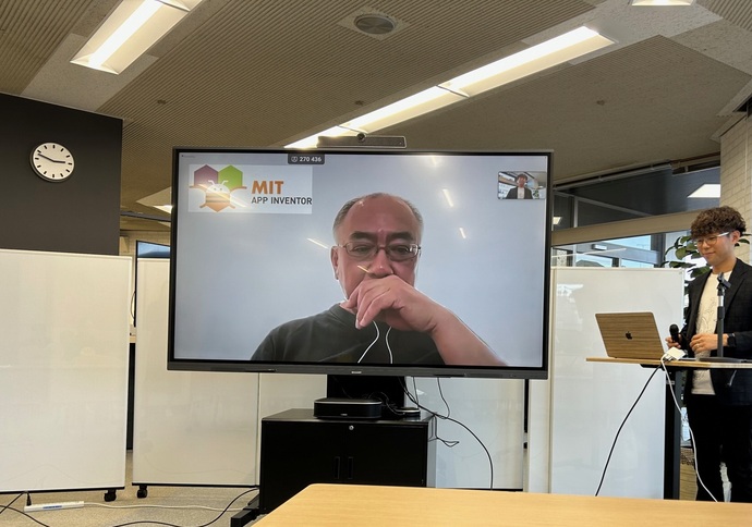 大型モニターにMIT App Inventorのロゴとオンライン参加の石原先生が映し出され、その横でマイクを手にした若い男性が司会進行を行っているハイブリッド形式のイベント風景の写真