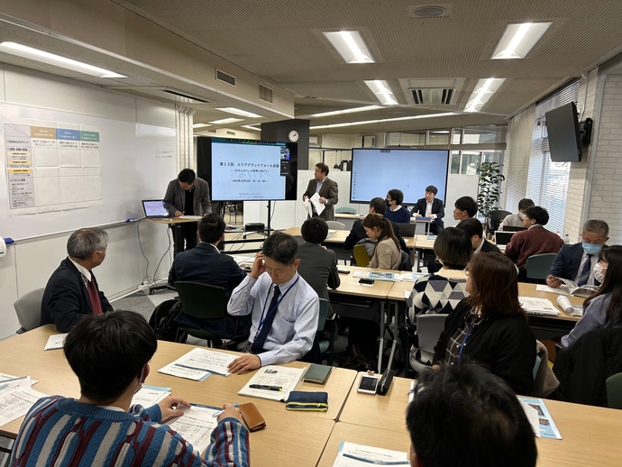 「第12回エリアプラットフォーム会議」と表示されたモニターの前で発表者が説明を行い、手前の長机に座った多くのスーツ姿の参加者たちが資料を見ながら熱心に耳を傾けている第12回会議の様子の写真