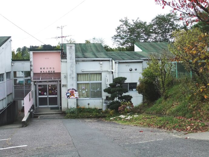 「高屋川市立 ぽえん保育園」という看板が掲げられた薄ピンク色と白の外壁の建物が、緑色の屋根と周囲の木々や斜面に囲まれて建っており、入口のガラス扉には花などの装飾が施されている、静かな雰囲気の建物の写真