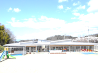 晴れ渡る青空の下、広々とした園庭の奥に平屋建てのモダンなこども園の校舎が建ち、左端にはカラフルな滑り台、テラスには遊具や荷物が並ぶ、明るく開放的な教育施設の全景を遠目から捉えた写真